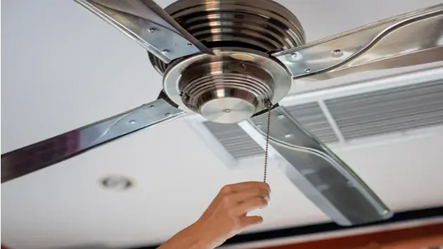 Can Ceiling Fans Reduce Wear On Your Air Conditioner? Person reaching up to turn on the ceiling fan, with an AC vent visible in the background.